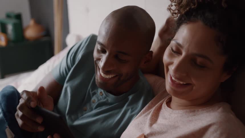 A person is lying on the couch in a living room looking at their phone another person is sitting next to them and pointing at the phone making both of them laugh