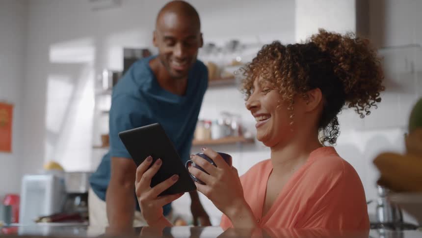 A woman is sitting at a counter looking at a tablet with a man standing behind her she is holding a mug and laughing while the man smiles as he points to the tablet