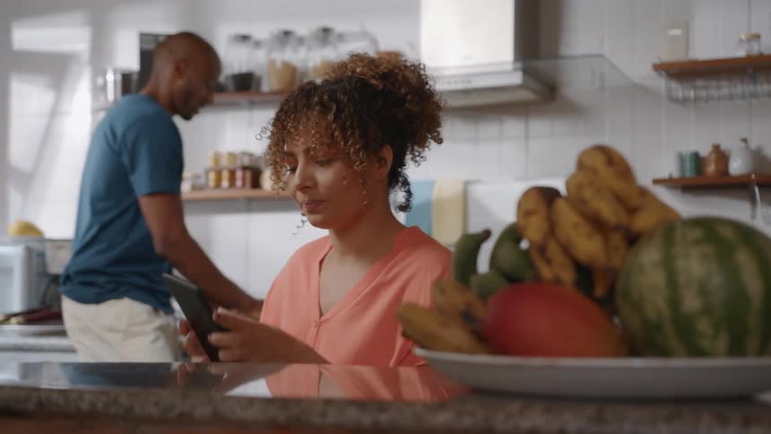 A couple is in the kitchen at midday the man is preparing a meal while the woman sitting nearby with a bowl of fruit beside her drinks coffee and scrolls through her phone