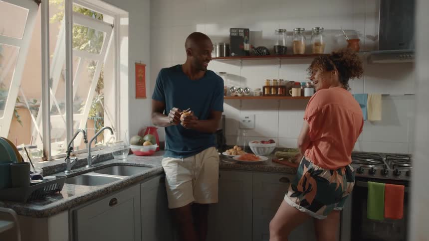 A happy couple stands together in a kitchen with a man in a blue tshirt and white shorts peeling fruit and talking to a woman in an orange tshirt who is preparing vegetables at the counter