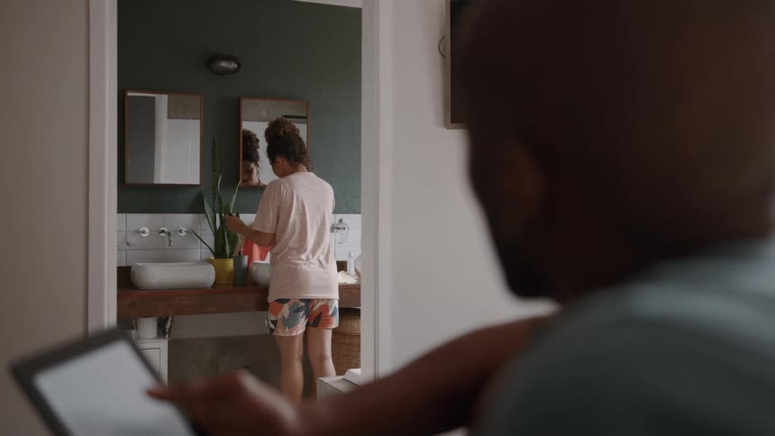 A woman is standing in a bathroom adjusting a houseplant a man is sitting on a bed in a separate room looking at a tablet