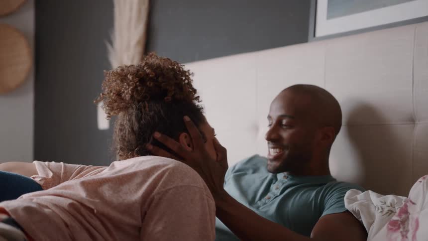 A couple is lying in bed talking and smiling the woman is on her side leaning on her elbow and the man is lying on his back