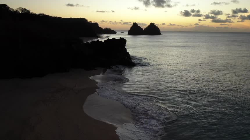 An aerial shot lowers over a silhouetted coastline at sunrise capturing waves gently washing onto the sand with two large rock formations in the distant ocean