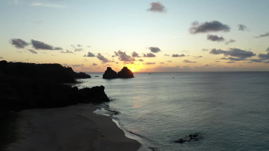 Flying through an opening in silhouetted tree branches the camera reveals a stunning sunrise over the ocean a dark coastline and two prominent rock formations