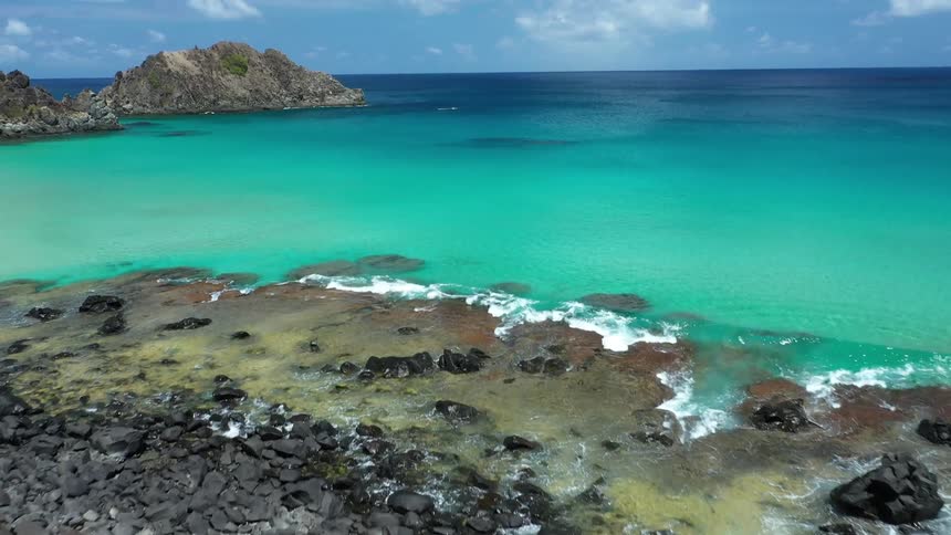 A beautiful drone shot pans to reveal a secluded beach with black rocks and lush green trees next to a large ocean of turquoise water with a rocky island in the distance
