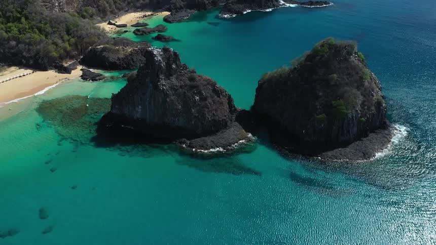 An aerial shot captures two massive rock formations sitting in the vibrant turquoise ocean just off a sandy beach backed by green cliffs
