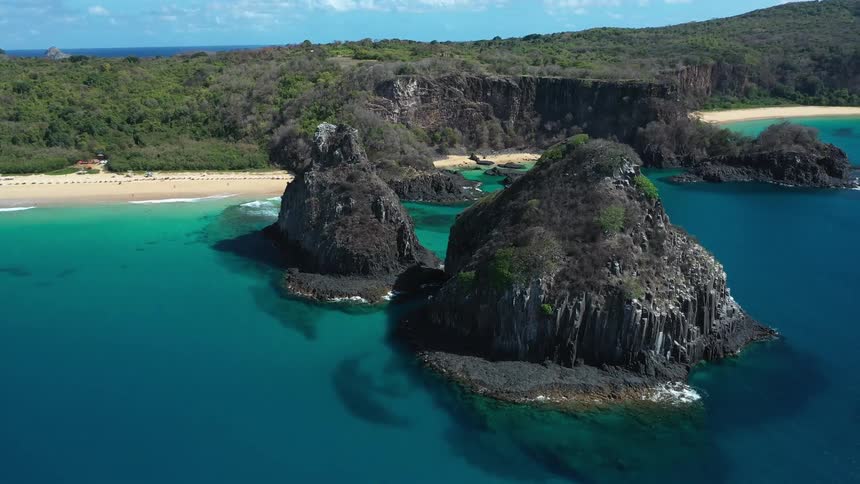 An aerial shot shows two large rock formations known as sea stacks rising from a turquoise ocean bay in front of a sandy beach and green hills