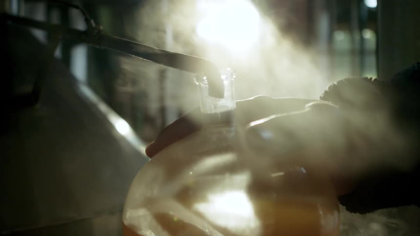 A persons hands are holding a round glass flask filled with an ambercolored liquid with a tube directed into the flask and steam rising into the bright sunlit air filled with dust