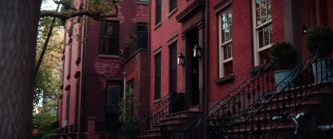 A shot of a street lined with red brick townhouses is shown with a tree in the foreground the camera pans from left to right showing the details of the buildings
