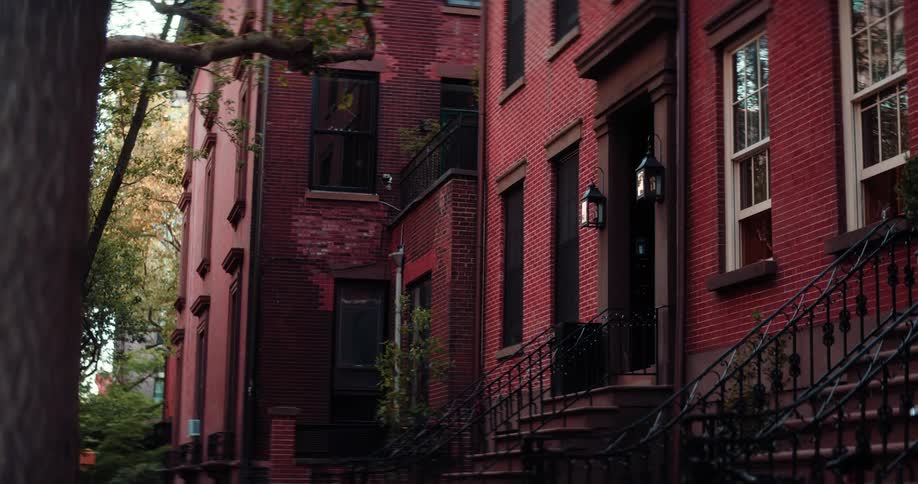 A shot of a street lined with red brick townhouses is shown with a tree in the foreground the camera pans from left to right showing the details of the buildings
