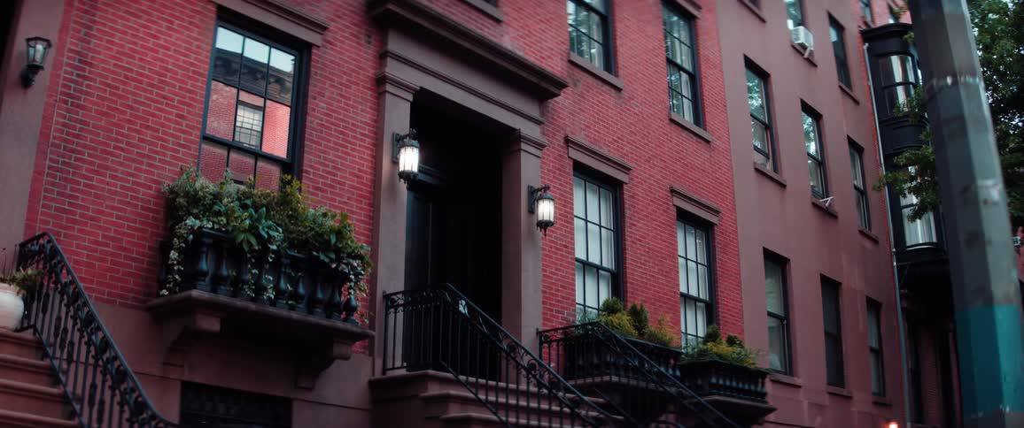 A shot of a fourstory brick apartment building is shown the facade is red brick with black railings and window frames