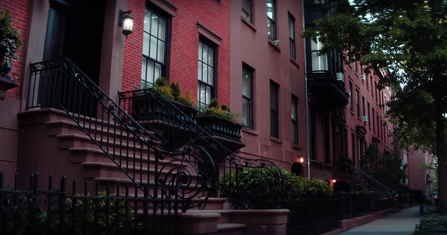 A view of a classic city street reveals a row of red brick townhouses with their front stoops ornate iron railings and glowing lanterns