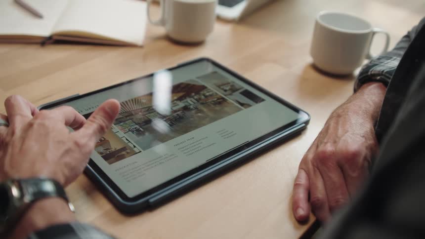 A closeup shot of a persons hands and forearms scrolling and tapping on a tablet while an open notebook and mugs sit on a table nearby