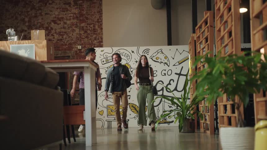 A woman in a brown top and two men in casual clothes are walking and smiling together through a modern office hallway with exposed brick walls and a large mural that reads we go together