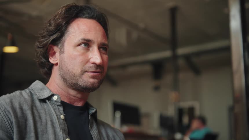 A shot of a middleaged man with dark wavy hair and a beard wearing a grey shirt he is looking offcamera and has a serious or thoughtful expression on his face