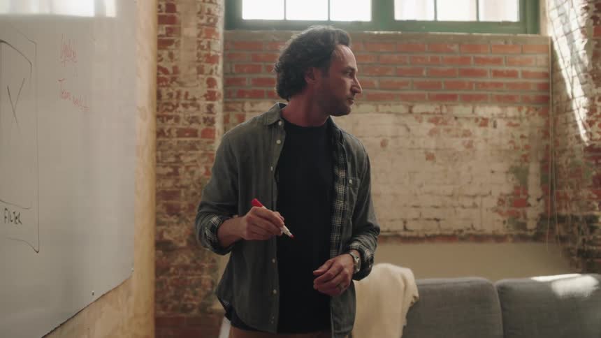 A closeup of a man with brown curly hair and a beard wearing a grey shirt over a black tshirt he is standing and drawing on a white board with a red marker