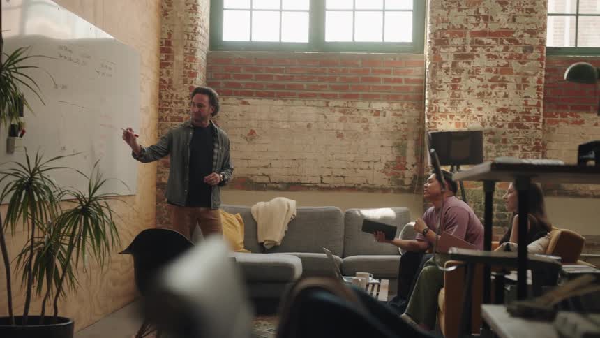A shot of a man standing and talking in a modern office space he is gesturing with his hands while addressing a small group of people who are sitting on a couch