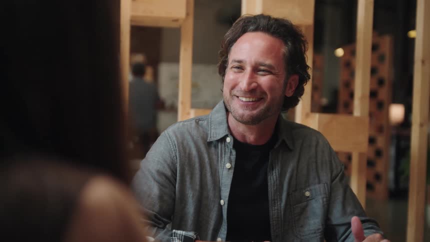 A medium closeup shot of a man in a modern office space smiling and talking he is sitting at a desk with a woman and a laptop a tablet and two mugs are visible on the table