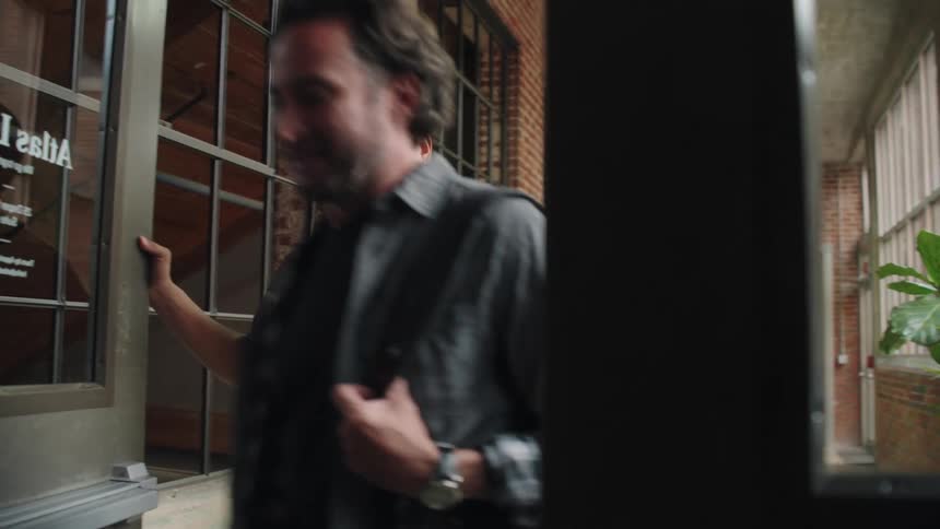 A wide shot shows three people walking in a hallway with large windows and brick walls the person in front opens a door for the other two people to pass through