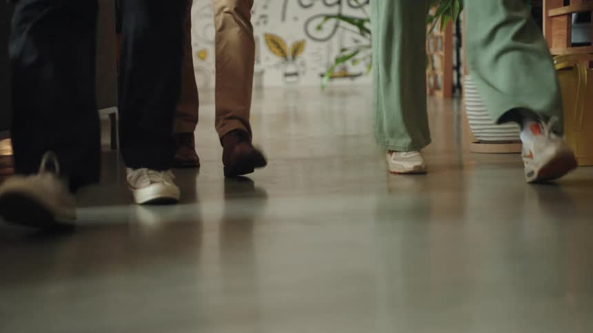 A shot from below shows the feet of three people walking they are walking on a smooth grey floor with a plant and yellow pot visible in the background