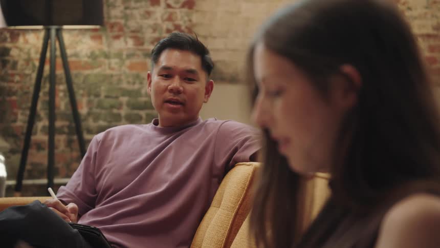 A smiling man in a pink tshirt is sitting on a couch and talking to a woman while taking notes