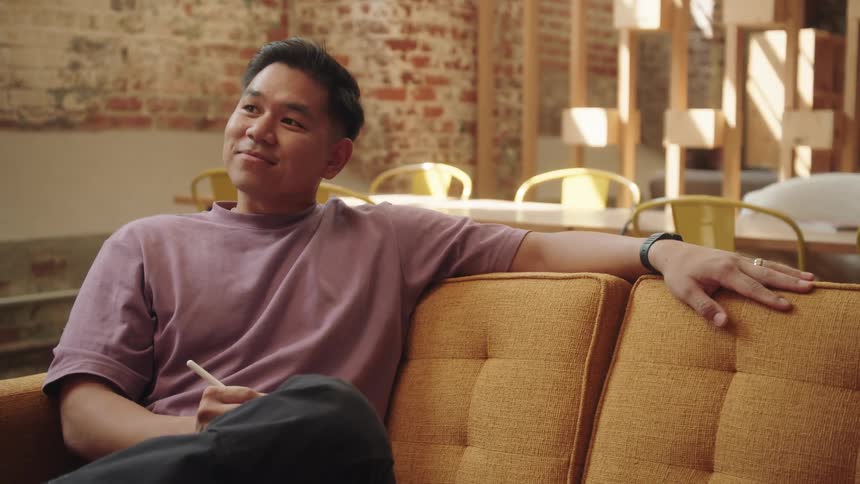 A medium shot of an asian man is shown as he sits on a yellow couch he is wearing a purple shirt resting his left arm on the back of the couch and holding a pen in his right hand