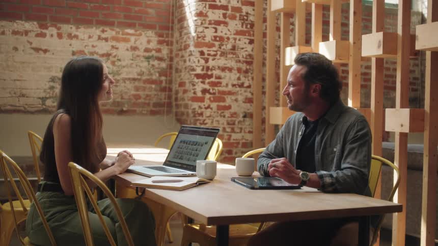 A man and woman are sitting at a table in a modern rusticstyle office they are talking and using a laptop and tablet seemingly collaborating on a project