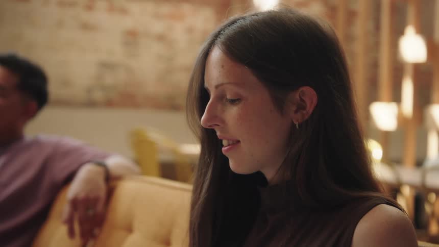 A smiling woman with long brown hair is listening to a lecture as she sits on a mustard yellow couch in a loft style room