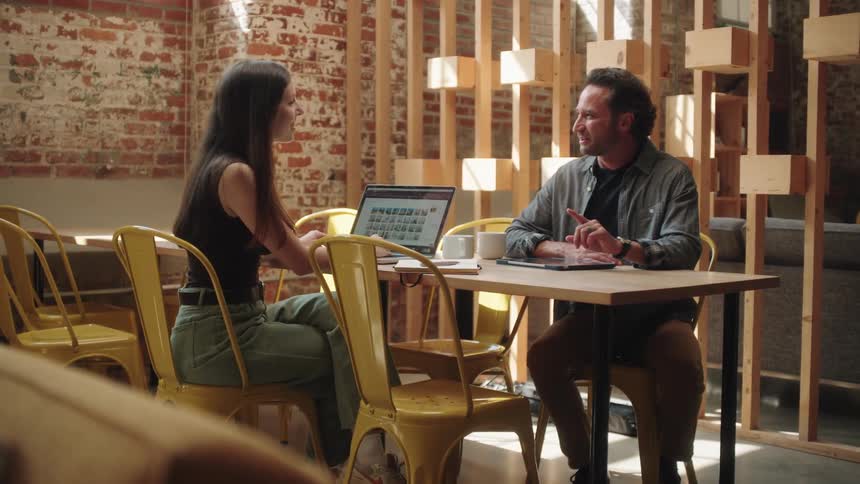 Man and woman sitting at a table with a laptop and a tablet have a conversation