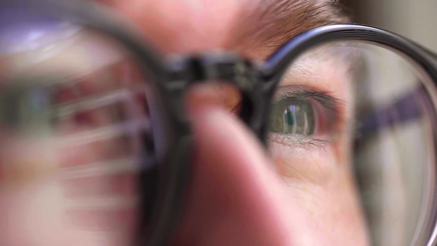 A man is shown in a very closeup shot with his eyes behind a pair of glasses the light from a screen is reflected in the lenses of his glasses