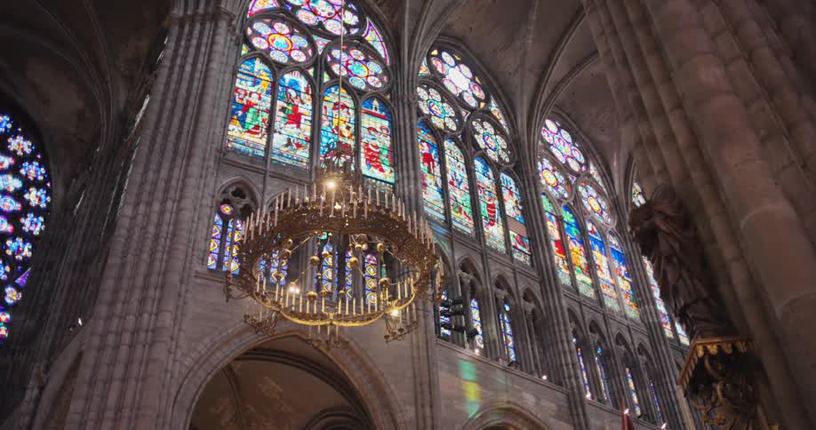The video shows a wide shot of the interior of a grand cathedral featuring magnificent stainedglass windows that line the high walls a large ornate chandelier hangs from the vaulted ceiling