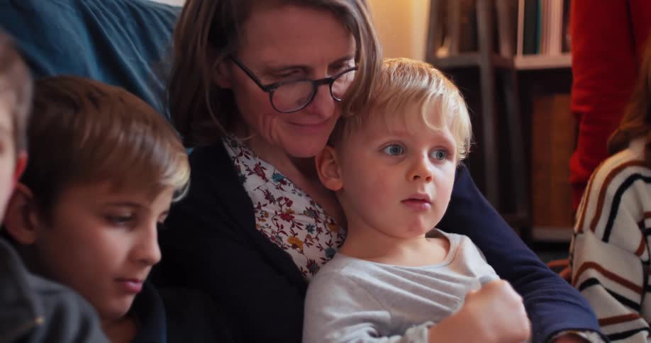 A mother with glasses is holding her young blonde son in her lap the boy looks forward with a wideeyed expression another child is visible on the left side of the frame
