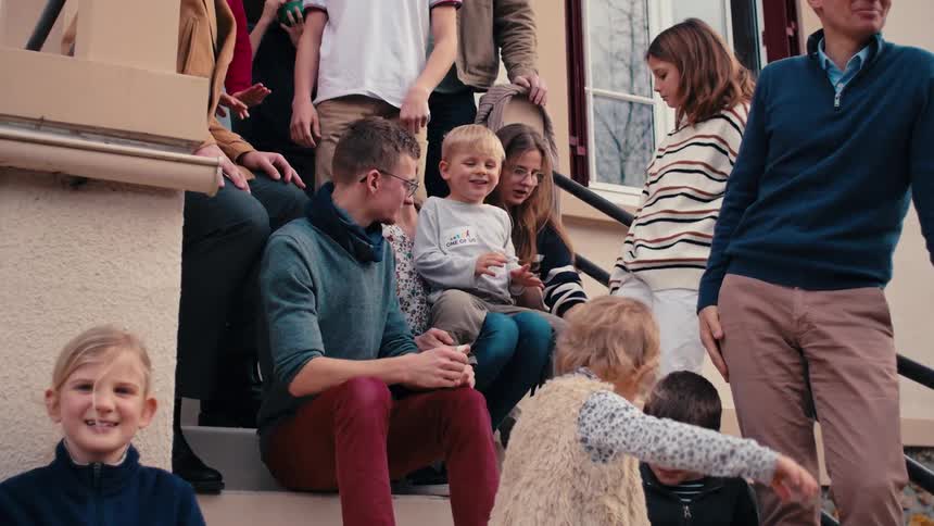 A casual shot of a large family of varying ages gathered and interacting on the stone steps of a building with children playing and others engaged in conversation
