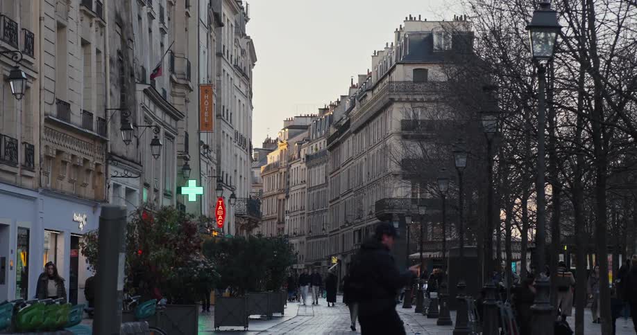 A panoramic shot captures the facade of some buildings with people walking on the sidewalk and on the streets