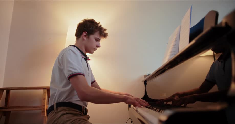 A young man sits and plays a grand piano indoors with a glowing lamp beside him