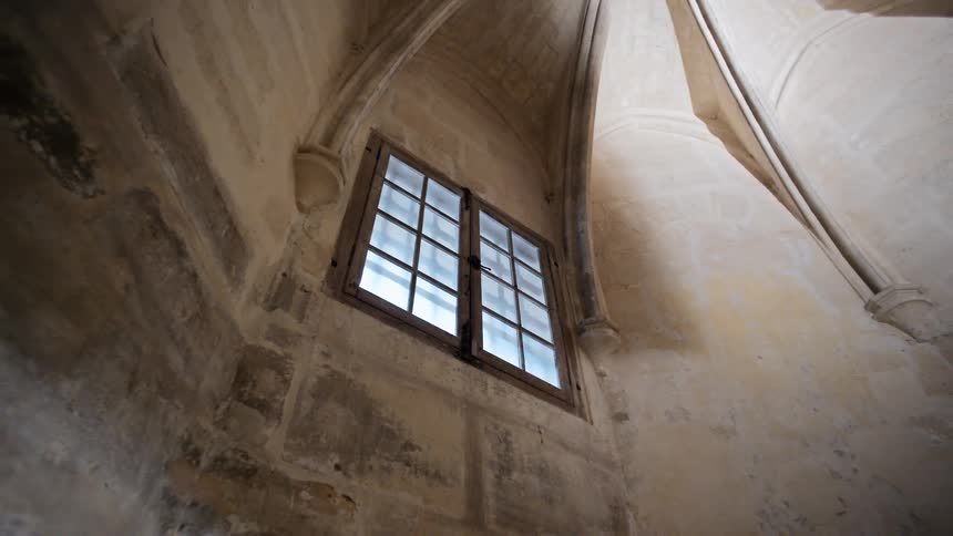 The video shows the interior of a historic building with stone walls and a vaulted ceiling a large gridpaned window is set high in the wall with light streaming in from outside