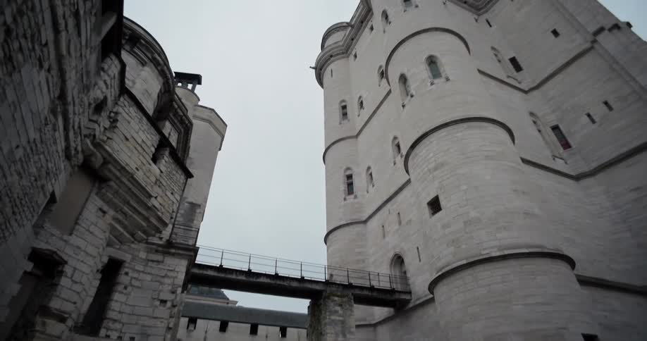 A large old stone fortress with a crumbling wall and a tall round tower are connected by a metal bridge in a stone courtyard on a gloomy overcast day