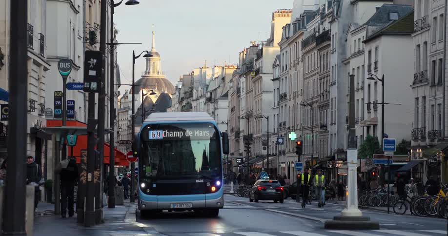 A street in paris is bustling with activity as a bus labeled 69 champ de mars drives towards the viewer pedestrians walk on the sidewalk and a car and other buses can be seen in the background