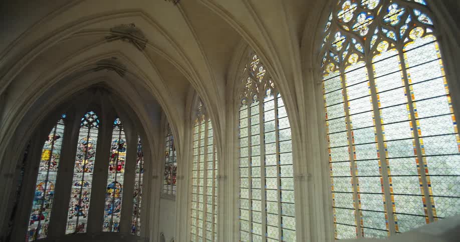 The video shows a wide shot of a cathedrals interior featuring a series of tall arched windows sunlight streams through the intricately designed stained glass illuminating the high vaulted ceilings