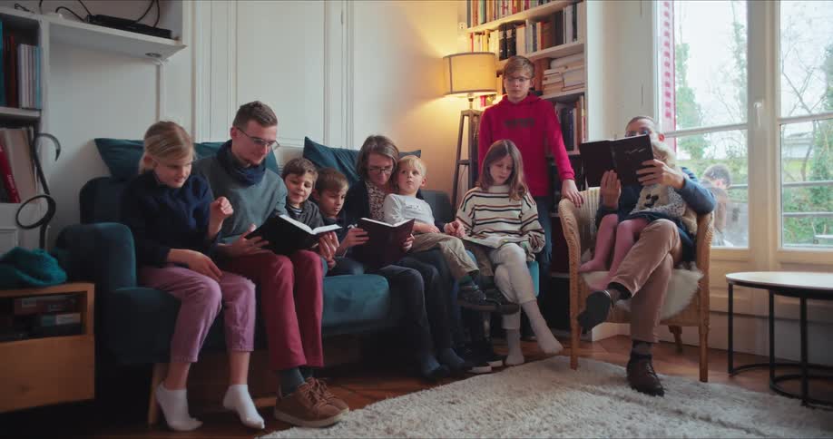 A family consisting of a father mother and several kids all seated on a couch in the home library reading together at midday