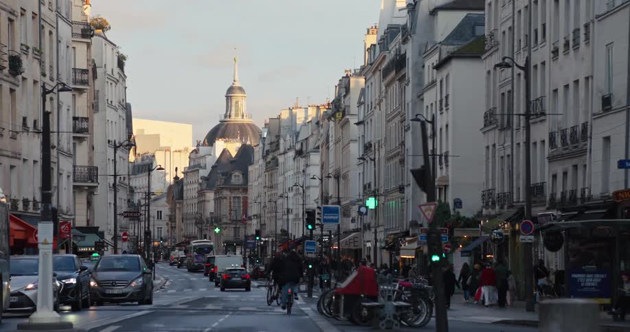 A long narrow european street is lined with tall white apartment buildings and filled with cars and people on bicycles with a large domed building visible in the distance at the end of the road
