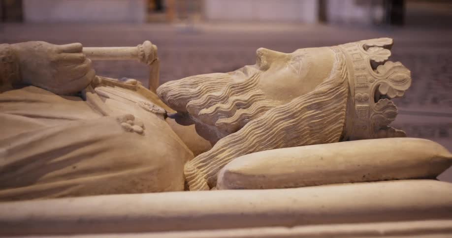 The video shows a closeup of a carved stone effigy of a king or nobleman lying in repose the figure is depicted with a long wavy beard and is wearing an ornate crown a scepter or staff is clutched in its right hand