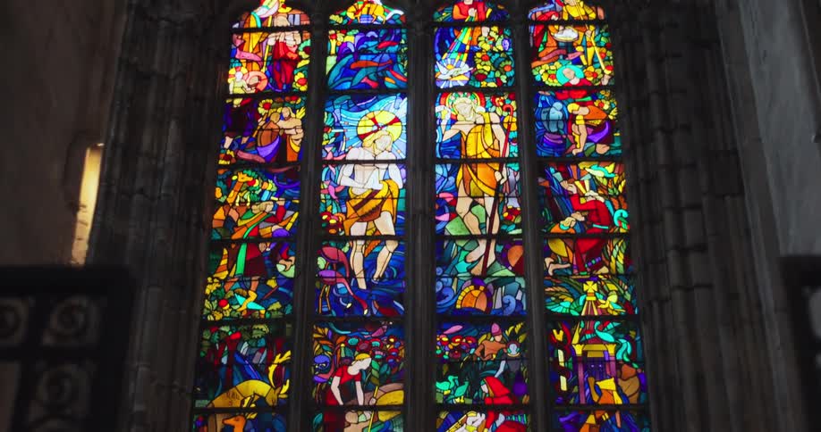 The interior of a grand cathedral with a detailed stained glass window depicting various religious scenes and a statue at the base of the window