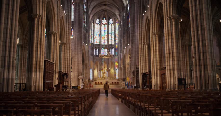 A lone person walks down the central aisle of a vast and ornate cathedral with towering columns and stained glass windows