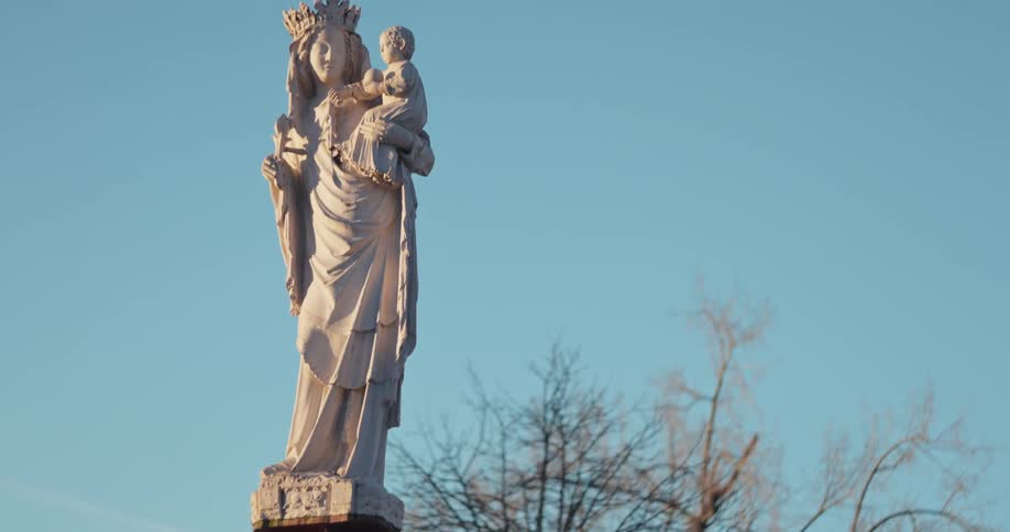 The video shows a statue of the virgin mary holding the baby jesus set against a clear blue sky the camera slowly pans up the statue revealing its full form and the detailed carvings of their crowns and robes