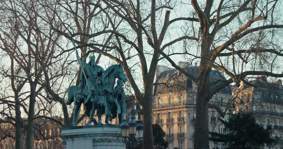 A shot of a bronze statue framed by the bare branches of trees with buildings on the background under a dusky sky