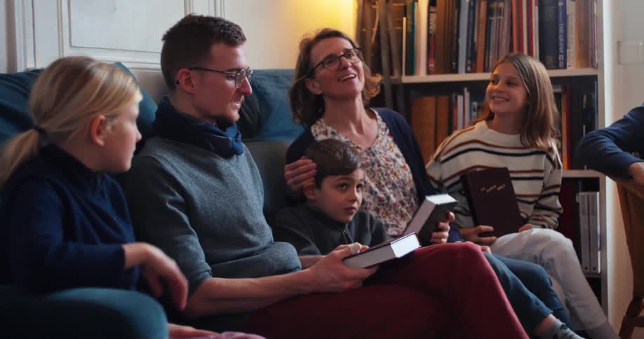A family with the mother walking in and sitting beside her son the father seated holding a book and the other kids sitting nearby all excited for a book reading in the home library at midday
