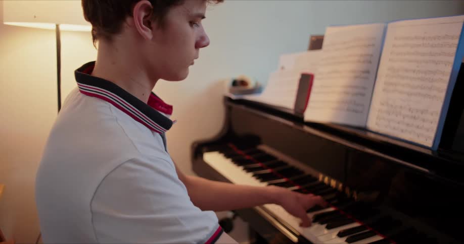 A teenage boy playing a grand piano with sheet music in front of him in his living room