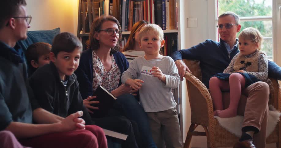 A large family including several children and adults is gathered in a living room they are seated together some holding books and appear to be engaged in a shared activity or conversation