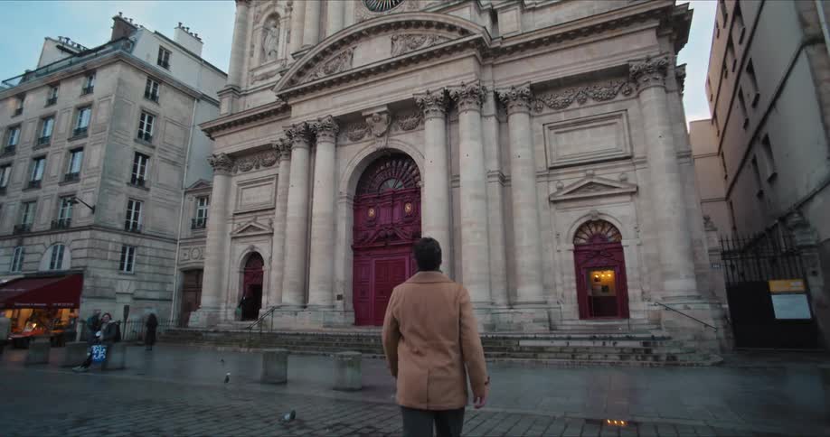 A man wearing a brown jacket walks toward a building with a clock tower looking up at the clock as he crosses the street at midday